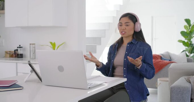 Asian adult woman starting work meeting on laptop at kitchen island wearing headphones taking notes