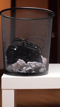 Vertical video old game controller dumped in a metal mesh wastebasket with crumpled paper, symbolizing e waste, obsolescence and digital detox