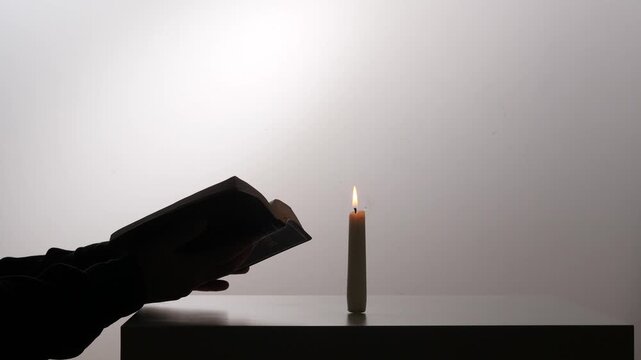 Silhouetted hands hold an open bible by candlelight, warm glow illuminating pages in a quiet moment of prayer, study and solemn spiritual reflection
