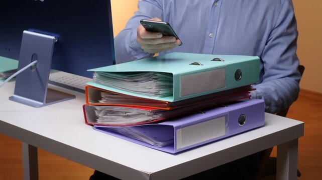 Office worker holding a smartphone behind a stack of document binders, organizing paperwork at a desk beside a computer, illustrating business administration and digital multitasking