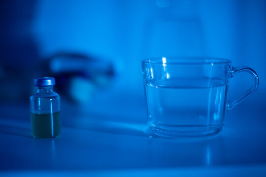 Glass cup and green vial glowing under blue LED light, sterile bench scene with shallow depth and grainy film texture, research sample staged for analysis, minimal clinical mood