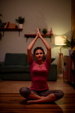 Woman seated cross-legged practicing overhead hand yoga pose in cozy living room with warm lamp light and calm focused expression at night, indoor mindfulness session