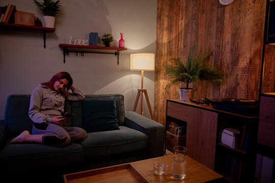 Woman slouched on teal sofa scrolling through phone in dimly lit apartment, soft lamp glow, wooden accent wall, potted plant, casual jacket, quiet introspective mood