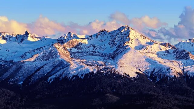 Snowy mountain peaks catch warm sunset light against a blue sky with fluffy clouds, capturing a majestic alpine vista.
