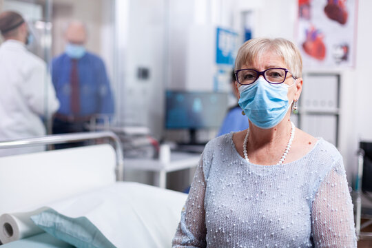 Senior, portrait and face mask with an older woman wearing glasses and a blue surgical mask beside a hospital bed, with medical staff behind her for clinical care and infection precautions.