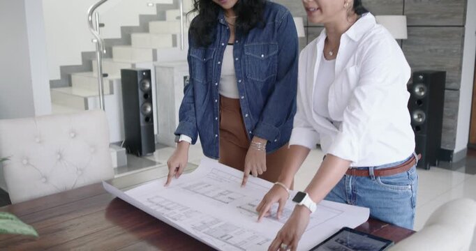 Indian women in white and denim unrolling plan at home, tracing rooms, tapping tablet for remodel