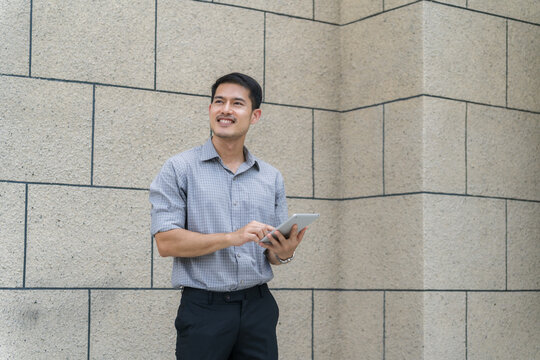 A confident Asian businessman in a smart casual shirt stands outdoor, smiling and holding a digital tablet while looking away against a stone wall background.