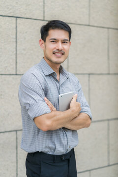 A confident Asian businessman in a smart casual shirt stands with crossed arms, smiling and holding a digital tablet against a stone wall background.