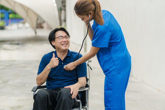 A female Asian nurse in blue scrubs checks a happy senior male patient in a wheelchair using a stethoscope outdoor.