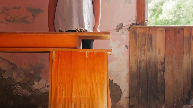 Woman wearing light t shirt leaning against wooden table in dilapidated room, faded coral walls, bright daylight streaming through large window, capturing solitude in deteriorating space