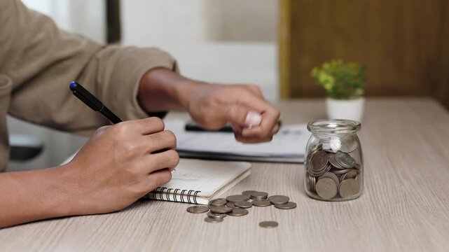 A person counts coins, puts money into a glass jar, and writes notes in a notebook on a wooden desk to manage personal finance, household budget, savings, and investments indoors at home.