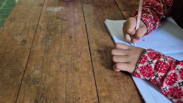 Close up of a student child writing in a notebook with a pencil wearing traditional batik school uniform at wooden desk