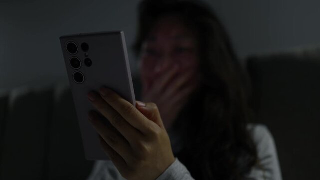 Frightened woman reading shocking news on smartphone at night. Asian woman lying on bed at home at night, holding her smartphone and reacting with shock and fear to something she reads on the screen.