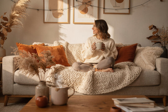 Cozy Autumn Living Room with Warm Textures and Natural Light . A comfortable living space with a knitted blanket, ceramic mug, and warm seasonal tones. woman in cosy clothes 