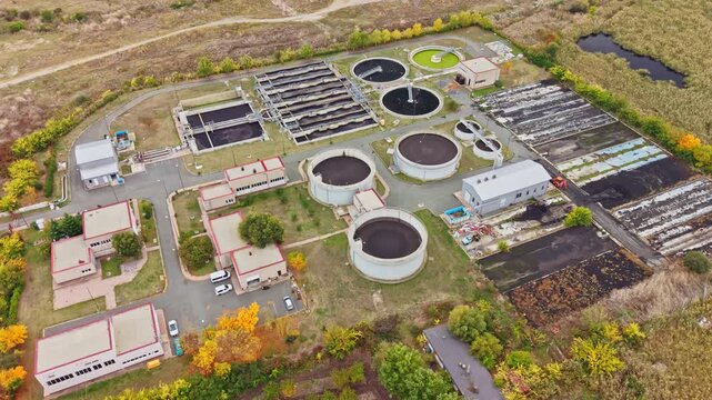 Circular settling tanks, treatment beds, service buildings, and access roads fill a wastewater treatment plant surrounded by fields and trees in daylight.