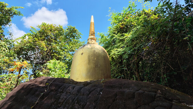 Golden stupa above dark stone base among tropical foliage under blue sky, creating serene Buddhist travel scene with vivid natural contrast and architectural detail