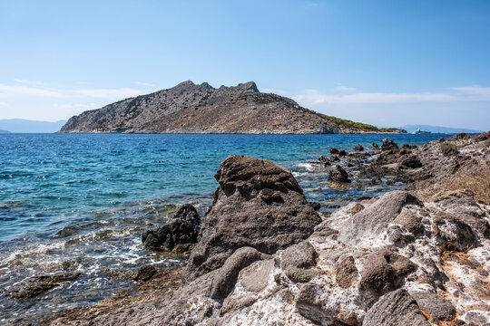 Clear turquoise waters lap against the rugged rocky shore near Perdika village on Aegina Island, offering a scenic view of Moni Island and distant sailboats in the Saronic Gulf.