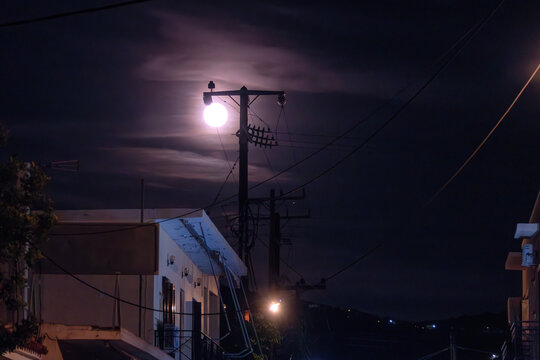 A bright full moon shines through wispy night clouds directly behind an electricity pole with power lines, illuminating the dark street and rooftops of a coastal village on Aegina Island, Greece.