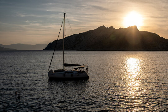 
A single sailboat rests in calm sea waters as the setting sun casts a golden glow and bright reflection across the surface behind a mountainous island coastline in Greece
