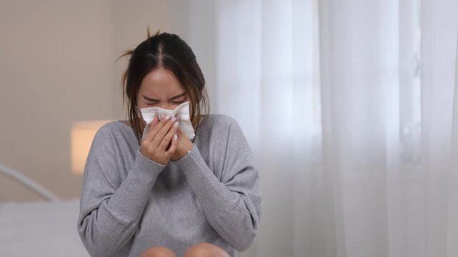 Sick Asian woman suffering from cold fever flu infection blowing runny nose with tissue napkin looking unwell exhausted at home bedroom