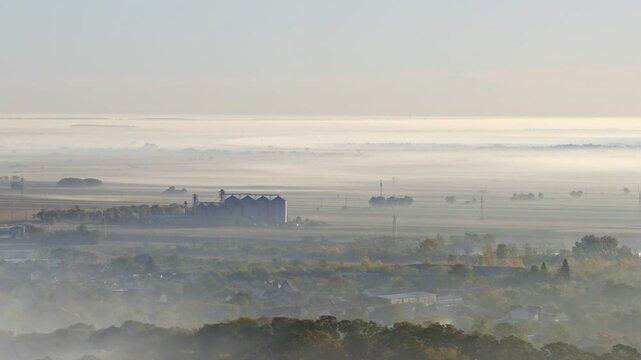 Aerial video across a shallow layer of ground fog and smoke drifting over a small farming community in serbian town of Temerin, Vojvodina. The sun sits low on the horizon at sunrise