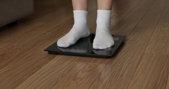 Woman feet in white socks stepping on digital electronic scales to check weight on wooden floor.