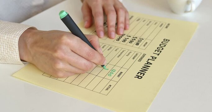 Woman hand writing actual expenses numbers with green marker on paper budget planner.