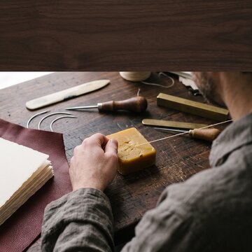 Artisan waxing thread with a block of beeswax for handmade leather bookbinding on a rustic wooden workbench surrounded by traditional tools