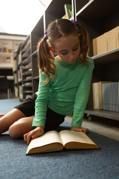 Schoolgirl sitting on floor and reading a book in library