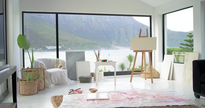 Easel holding blank canvas near paint table with brushes, drop cloth, windows showing misty lake