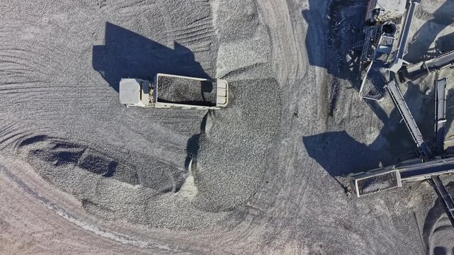 A loader moves gravel beside conveyor equipment at a work site during daylight, showing tracks in the aggregate and a career in construction.