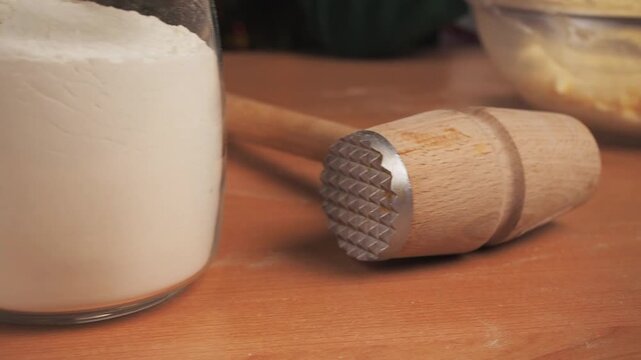 Close-up of wooden meat mallet with metal textured head on wooden table, jar of flour and bowl of dough blurred in background, kitchen tools and baking preparation