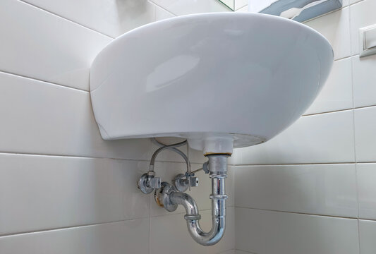 White ceramic bathroom sink with exposed chrome plumbing against tiled wall. Sink installation and drainage system. Residential plumbing fixture for hand washing and daily hygiene