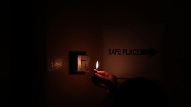 In a dark room, a woman holds a lit candle while pressing a light switch. A Safe Place sign with an arrow on the wall points toward a safe location. Power outage, lack of electricity, shelter, danger,