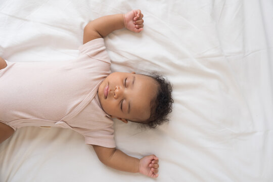 African American newborn baby sleeping soundly on white bed sheets with hands raised up, top view portrait of cute innocent infant napping comfortably in cozy bedroom