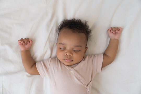 African American newborn baby sleeping soundly on white bed sheets with hands raised up, top view portrait of cute innocent infant napping comfortably in cozy bedroom