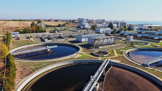Circular wastewater treatment basins, pipes, and processing buildings sit among fields on the outskirts of a town under a clear daytime sky.