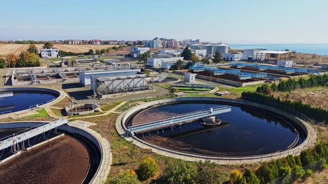 Circular treatment tanks, pipes, buildings, and service roads fill a coastal wastewater plant during daytime, with the sea visible beyond the facility.