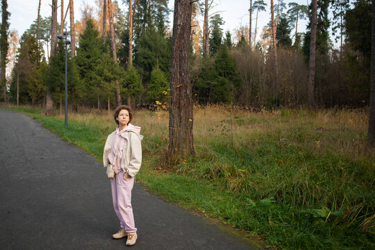 woman stands in casual pink clothing and sneakers on a curving path through a forest park, with her hands in her pockets and wide woodland space beside her for text