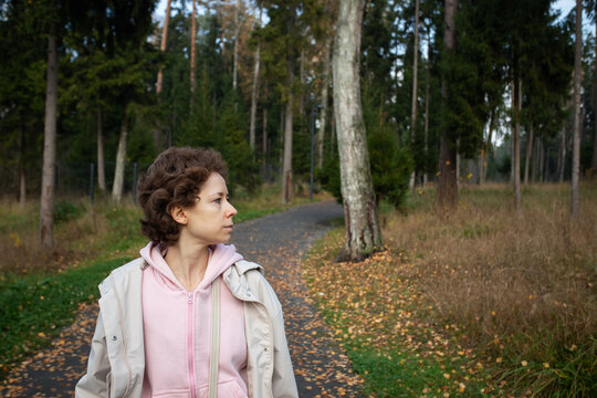 woman with short curly hair looks into an autumn woodland during a fresh air break, with her pink hoodie in the foreground and a curving path lined with fallen leaves behind