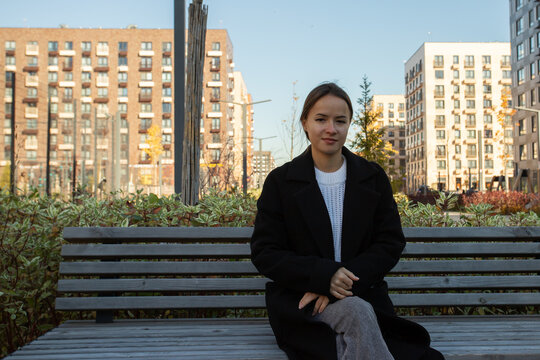 woman in a black coat sits with crossed legs on a wooden bench in a landscaped apartment courtyard, facing the camera with residential buildings and autumn shrubs behind her