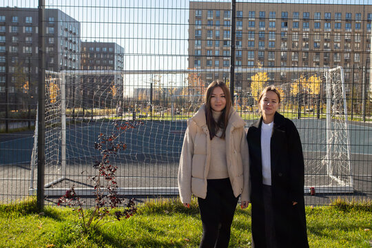 two women stand beside a fenced sports court in an apartment courtyard, with a soccer goal, sunlit grass and autumn trees around them, wearing a beige puffer vest and black coat