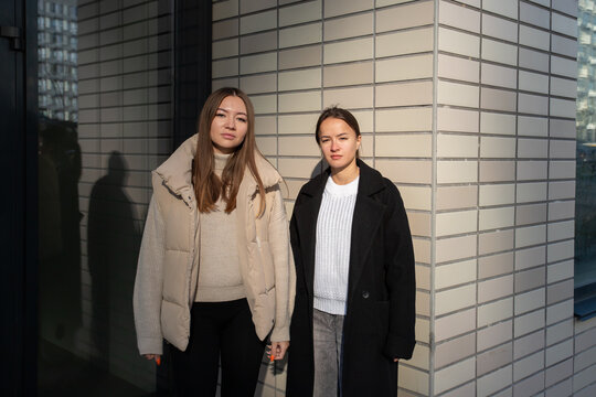 two women stand beside a gray brick facade in bright sunlight, wearing contrasting beige and black outerwear, with their silhouettes reflected in a dark glass door next to them