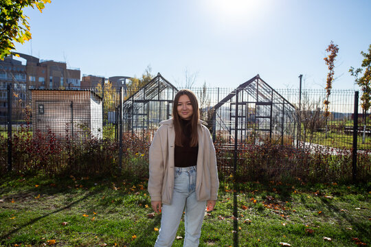 woman stands outside a fenced garden with two glass greenhouses, surrounded by autumn leaves and shrubs with apartment buildings nearby, facing the camera in bright sunlight