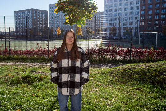 a woman with her hands in her pockets stands on grass near a fenced sports court, with a goal, autumn foliage and apartment buildings behind her, wearing a black and white plaid jacket