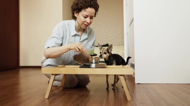 woman kneels behind a wooden table and scoops dry dog food from a storage container, with a bowl on a kitchen scale for portion control and a small dog peeking from behind