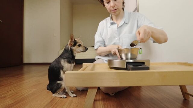 small dog stands beside a kneeling woman as she scoops dry food from a container, preparing a feeding portion with a metal bowl on a kitchen scale at a low wooden table