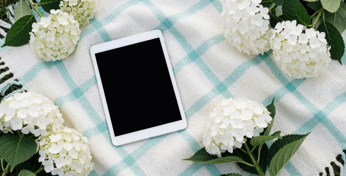Flat lay of digital tablet on white, turquoise plaid blanket. White hydrangea flowers surround electronic device on fabric. Minimalist workspace with floral decor for blog content, social media post,