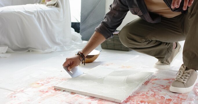 Indian man kneeling and spreading adhesive on tile with notched trowel near mixing bowl, copy space
