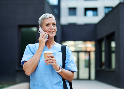 Nurse in scrubs enjoying coffee and using a mobile phone outside a modern medical facility during a sunny day